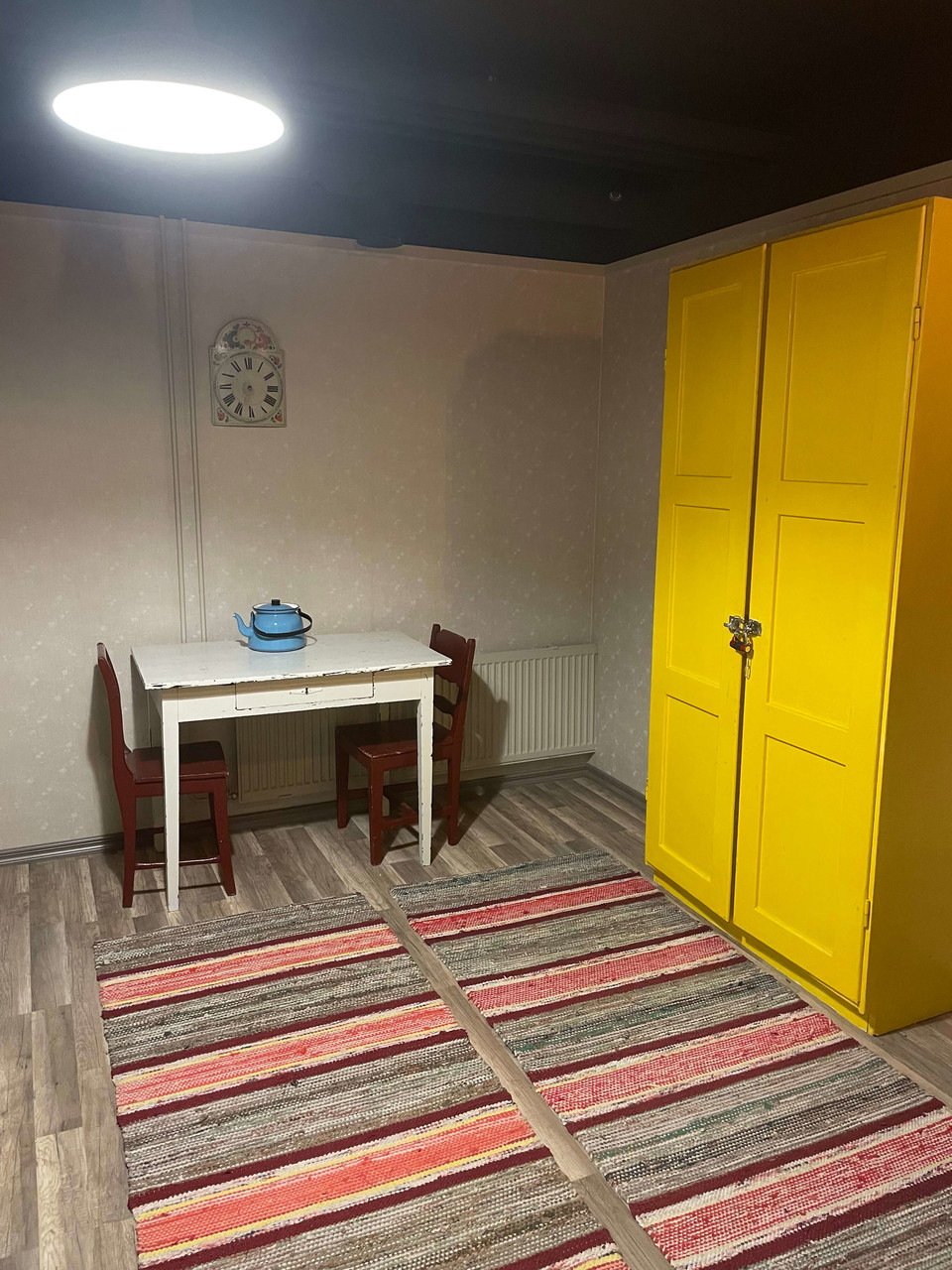  Syytön mies -huoneessa on keltainen kaapisto, kello seinällä, räsymattoja lattialla ja vanha valkoinen pöytä, jolla on sininen emalipannu. 