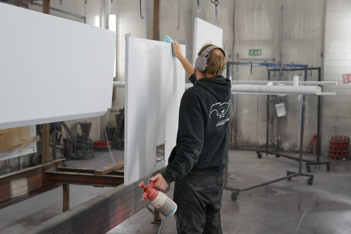 Mustaan huppariin ja housuihin pukeutunut työntekijä valmistelee säiliöauton luukkujen maalausta. Valkoiset luukut roikkuvat telineestä.