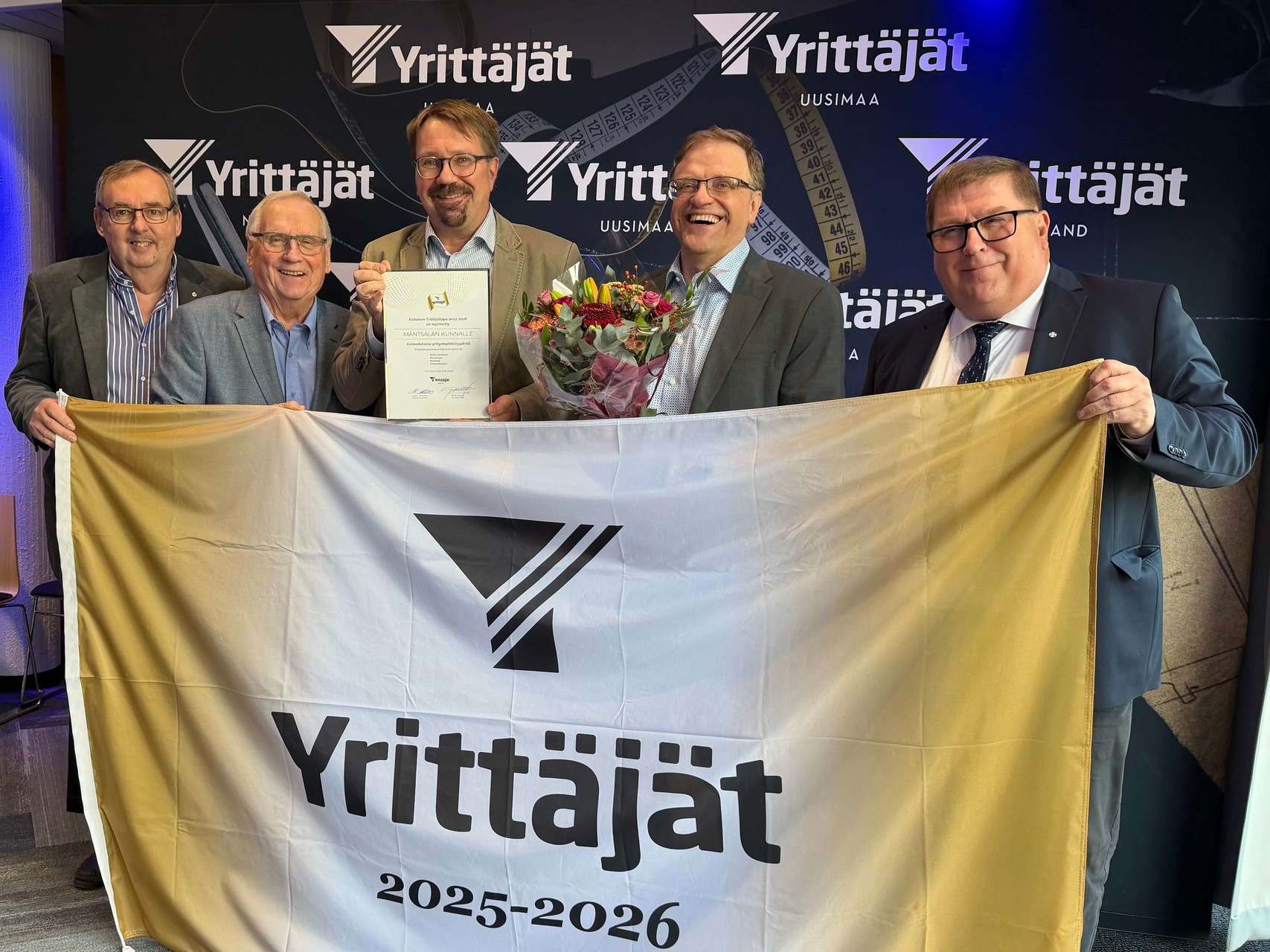 Kankaista kultaista yrittäjälippu yläreunasta pitelevät Mäntsälän Yrittäjien aiemmat puheenjohtajat Jarmo Mäkeläinen ja Pentti Sorkio, Mäntsälän Yrityskehityksen tj Juha Leinonen, kunnanjohtaja Hannu Laurila ja MY:n nykyinen pj Eero Kokko.   