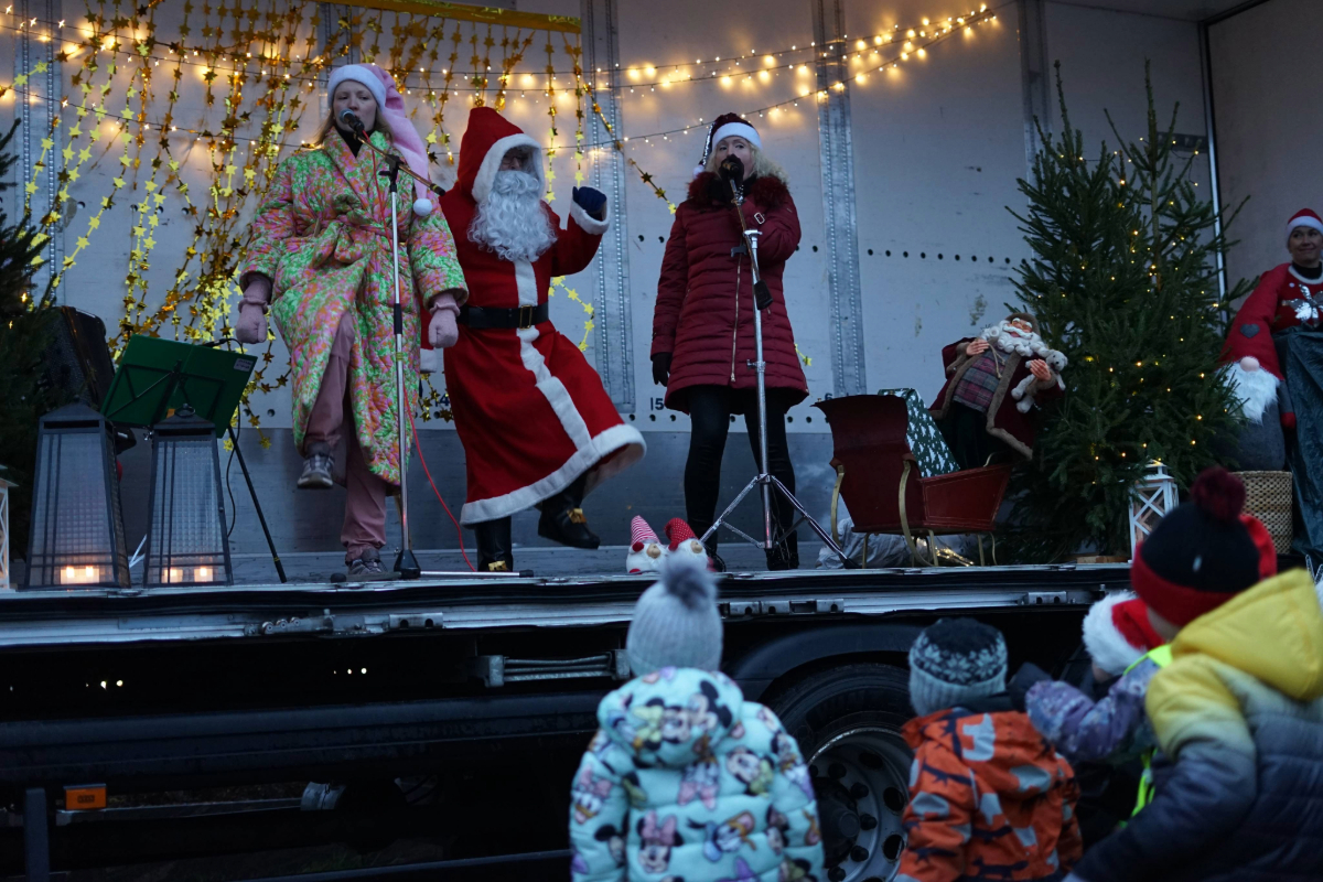 Joulupukki kuorma-auton sivusta avonaisella lavalla kahden naisen välissä. Lapset katsovat tanssiesitystä joulun tunnelmaan koristellulla lavalla.