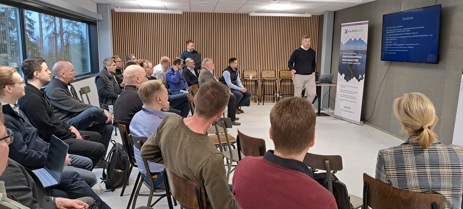 Salissa istuu ihmisiä puoliympyrään asetelluissa tuoliriveissä. Iiro Mikkola seisoo mainos roll-upin ja näytön vieressä.