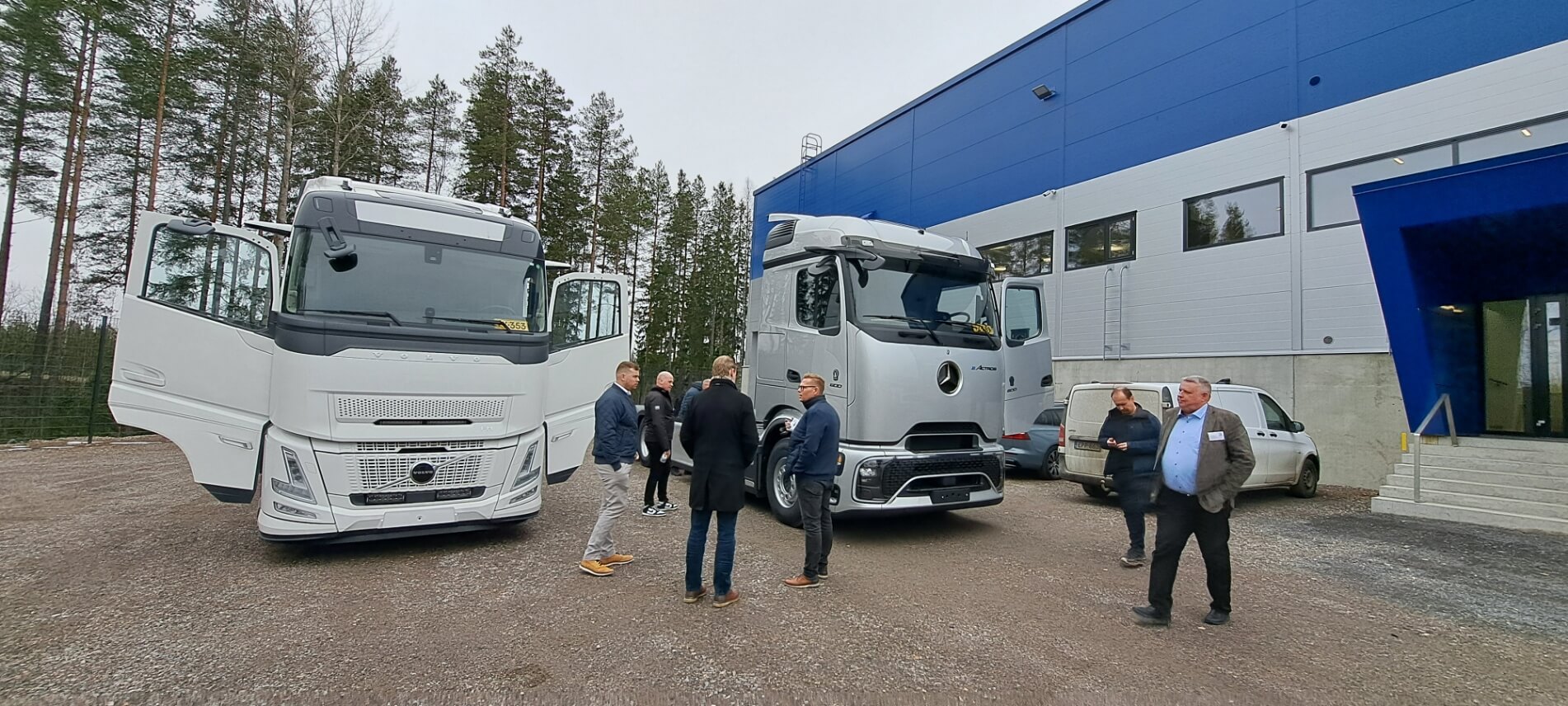 Miehiä juttelemassa kahden kuorma-auton edessä sini-harmaan logistiikkakeskuksen vieressä.