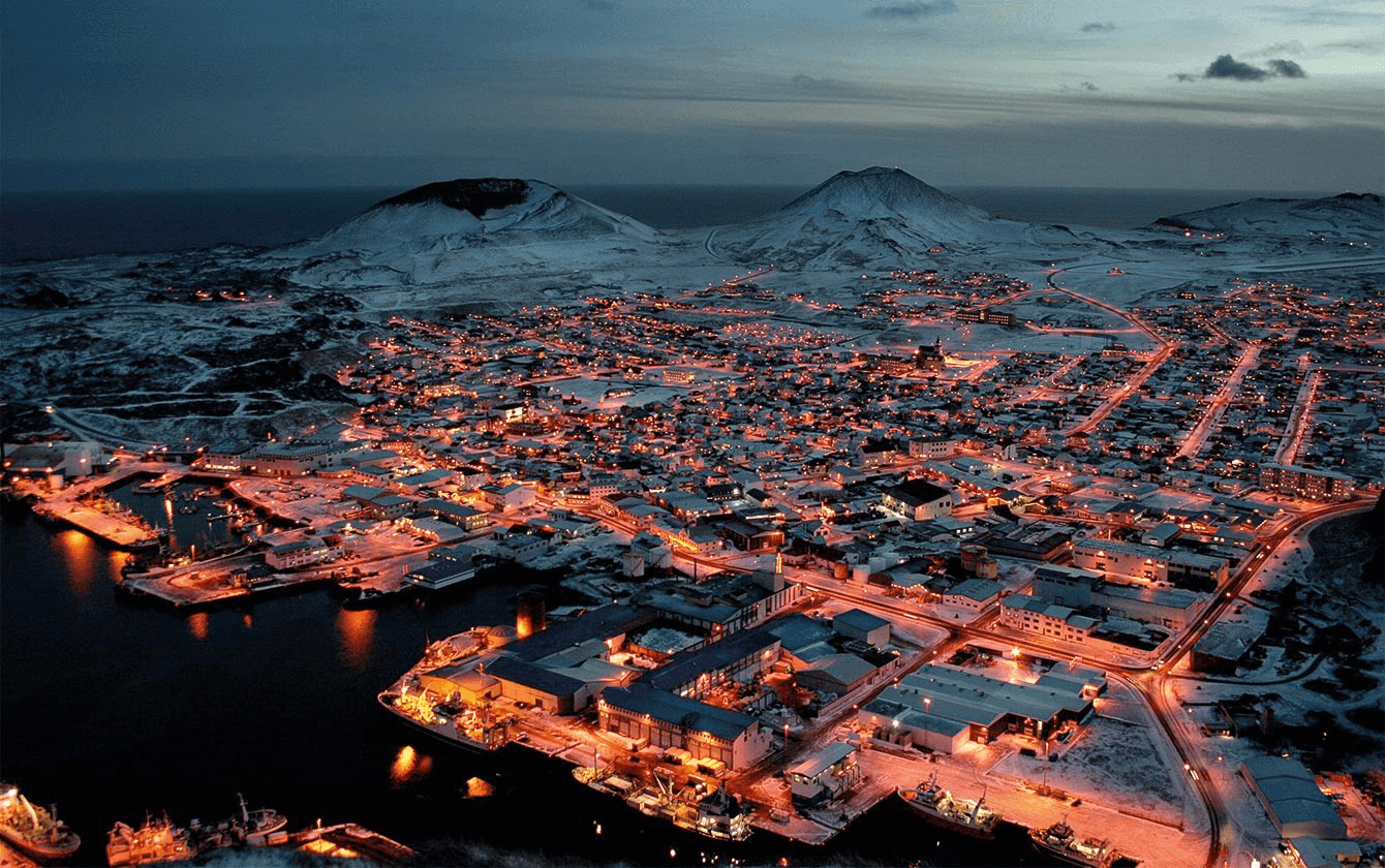 Islantilaisen Vestmannaeyjarin kylän keskustasta ilmakuva. Satamakylä on kuvattu satama-altaiden suunnalta pimeän aikaan niin että rakennukset ja tiet näkyvät valaistuina. Taustalla kohoaa vaaroja ja näkyy merta.