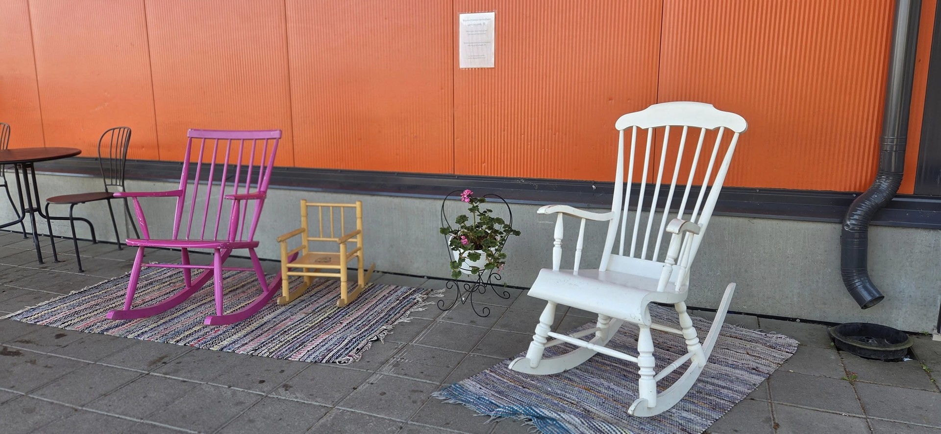 K-marketin oranssin seinustan vieressä värikkäillä räsymatoilla on erikokoisia ja värisiä keinutuoleja, pinkki, keltainen lasten tuoli ja valkoinen keinutuoli.