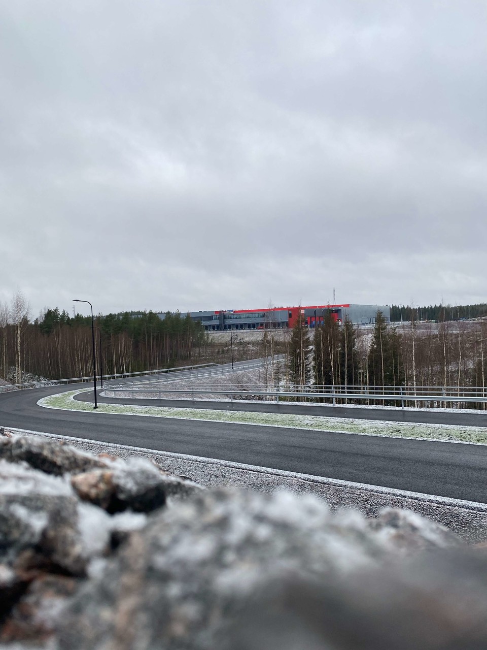 Tokmanni Groupin uusi  puna-harmaa Moreeni-logistiikkakeskus näkyy puuston takana ja asfalttitien päässä.