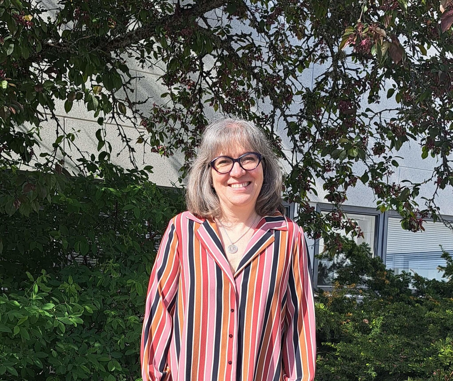 Hymyilevä Päivi Ahvenainen seisomassa puuston edessä yllään rento mustan ja eri punaisen sävyin raidoitettu paita ja tummasankaiset silmälasit.